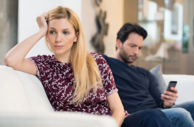 donna triste seduta sul divano e dietro di lei un uomo che guarda il cellulare